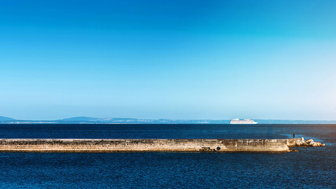 A single person stands on a long stone breakwater jutting out into the ocean with a large cruise ship in the distance, Estoril, Portugal, Europe