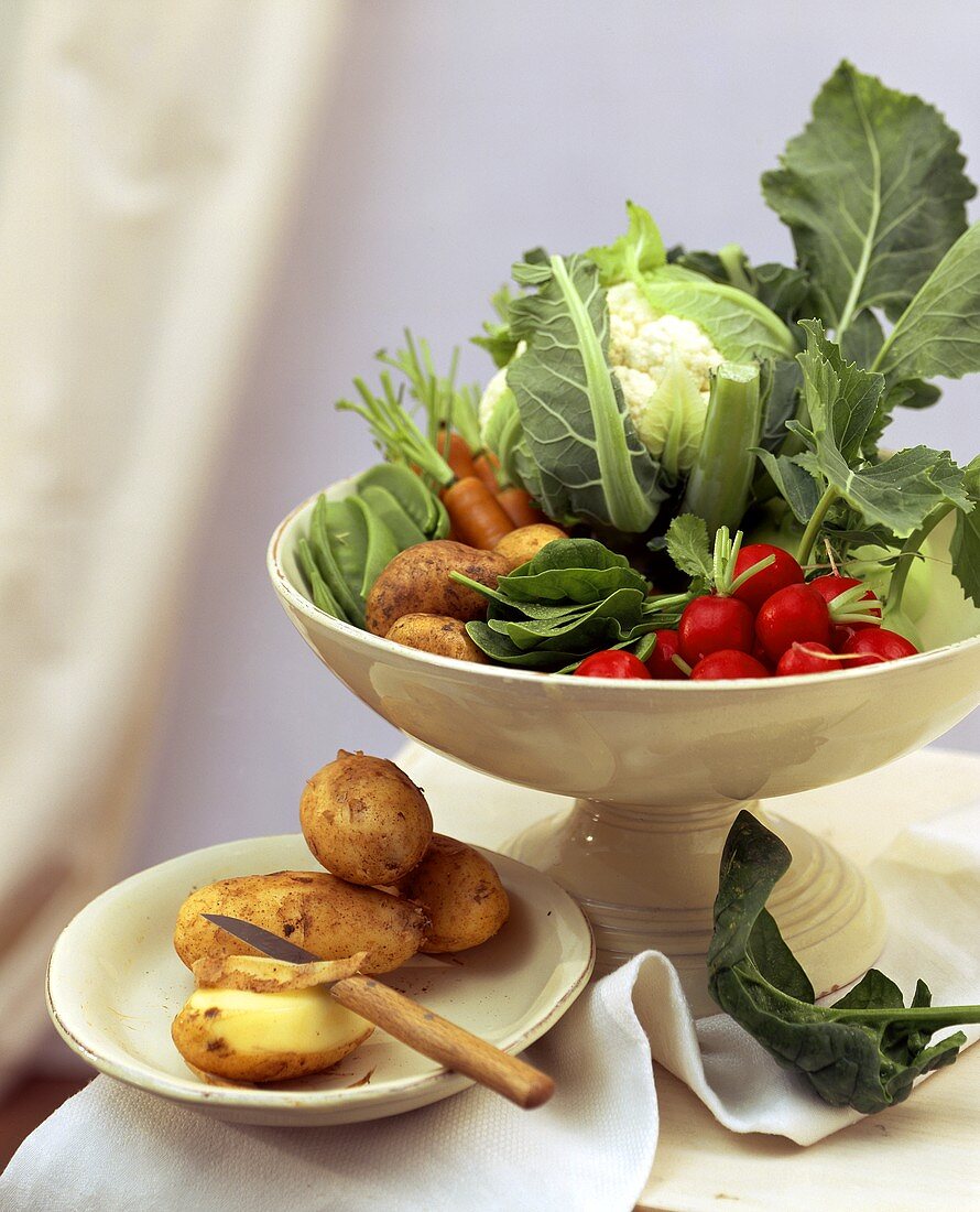 Vegetables in white bowl, potatoes on plate beside it