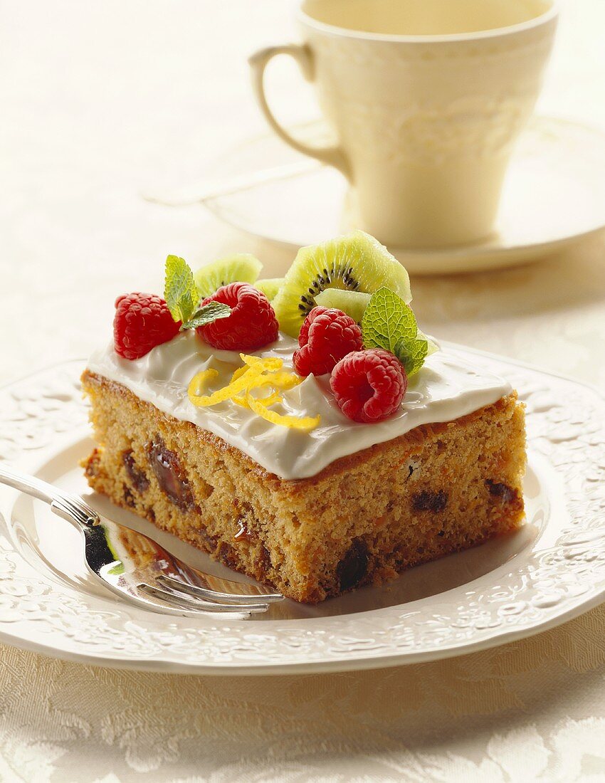 A piece of spice cake with white icing, raspberries & kiwi fruit