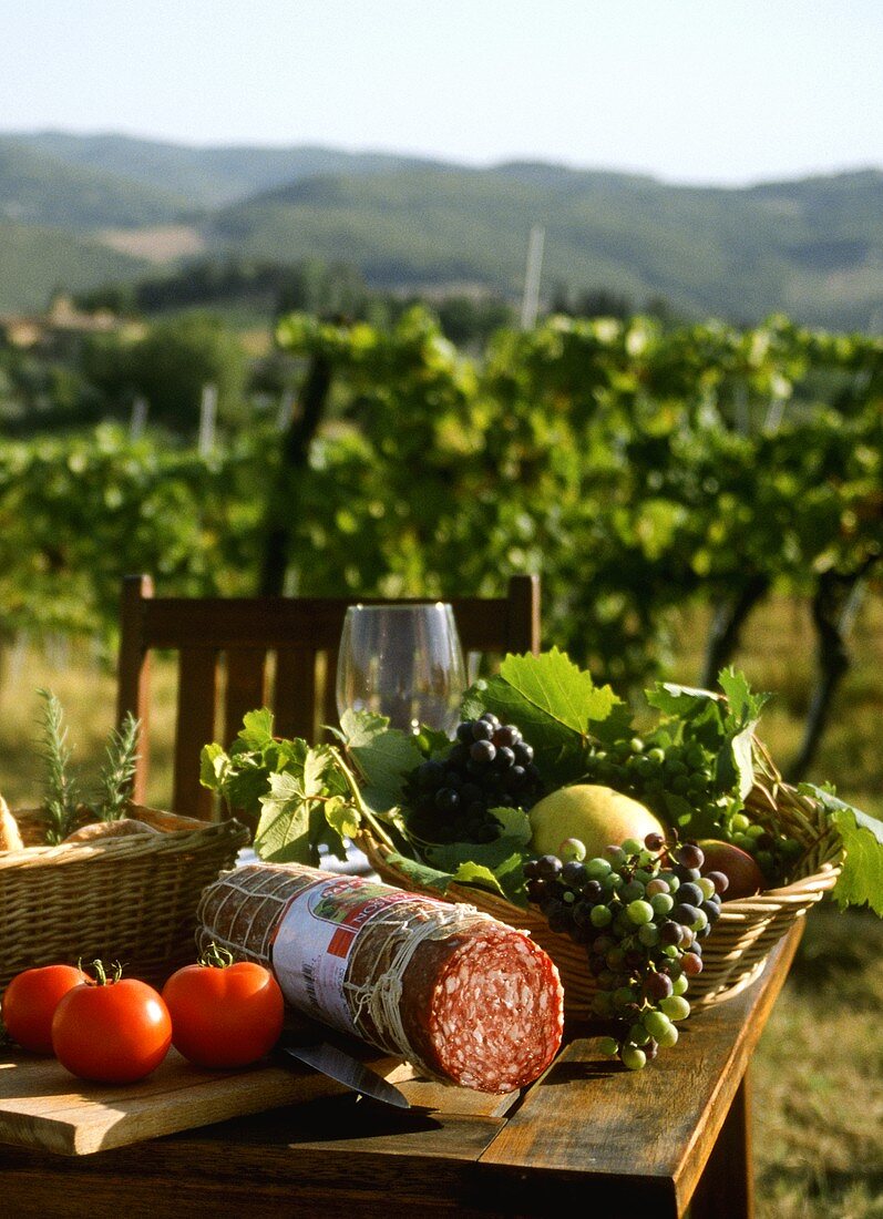 Table with salami, tomatoes, grapes in front of Tuscan vineyard