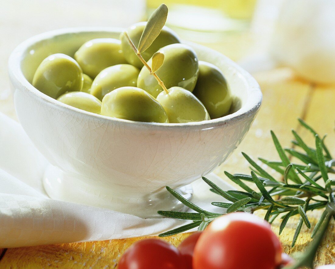 Grüne Oliven in einem Schälchen, daneben Rosmarin, Tomate