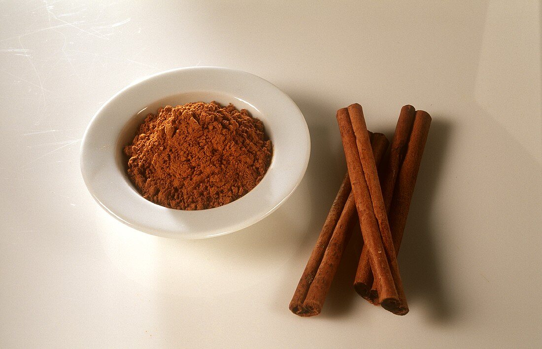Cinnamon powder in white bowl, cinnamon sticks beside it
