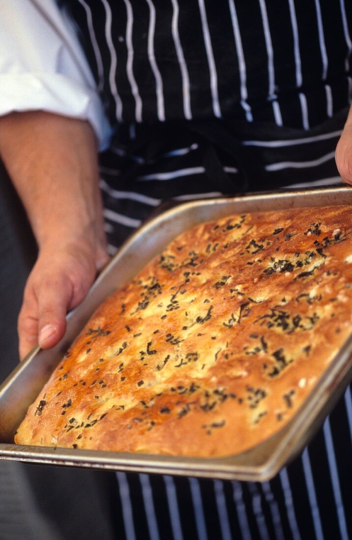 Person holding baking tray with herb focaccia (Ital. Flatbread)