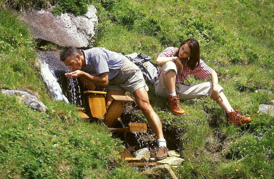 Junges Paar in den Bergen, Mann trinkt Wasser aus Bächlein