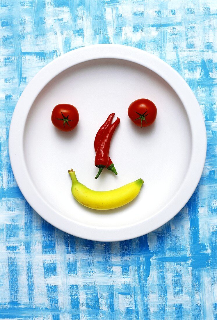 Food collage: face made from banana, chili & tomatoes on plate