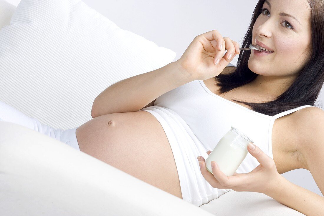 Schwangere Frau auf Sofa liegend, mit Glas Naturjoghurt
