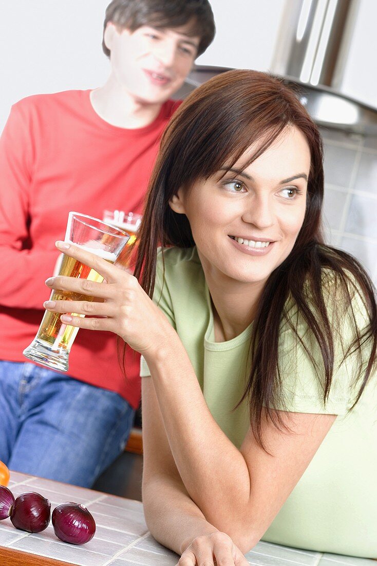 Young woman in kitchen with beer glass, man in background