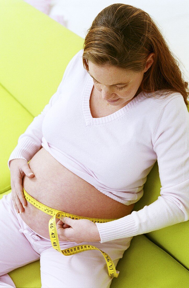 Schwangere Frau auf dem Sofa hält Massband um den Bauch