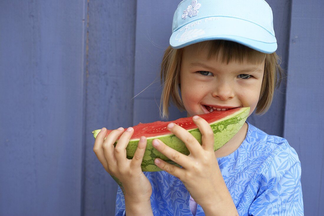 Mädchen mit Melonenschnitz im Mund vor blauer Wand