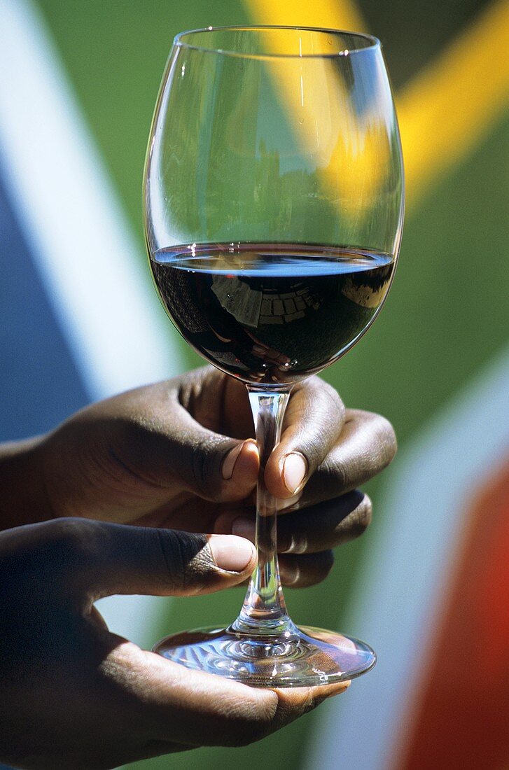 Hand hält ein Glas Rotwein vor südafrikanischer Fahne