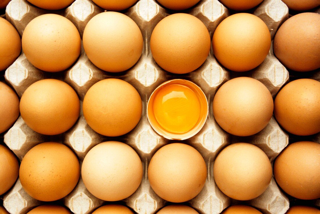 Braune Eier im Eierkarton, darunter ein aufgeschlagenes Ei
