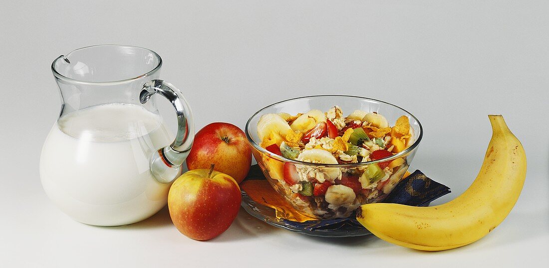 Müsli mit Früchten in Glasschale; Milchkrug, Äpfel und Banane