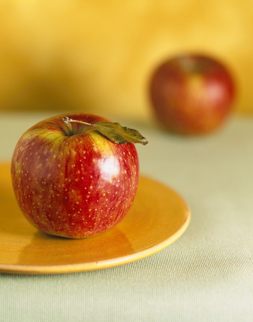 Red apple with leaf on yellow plate in front of second apple