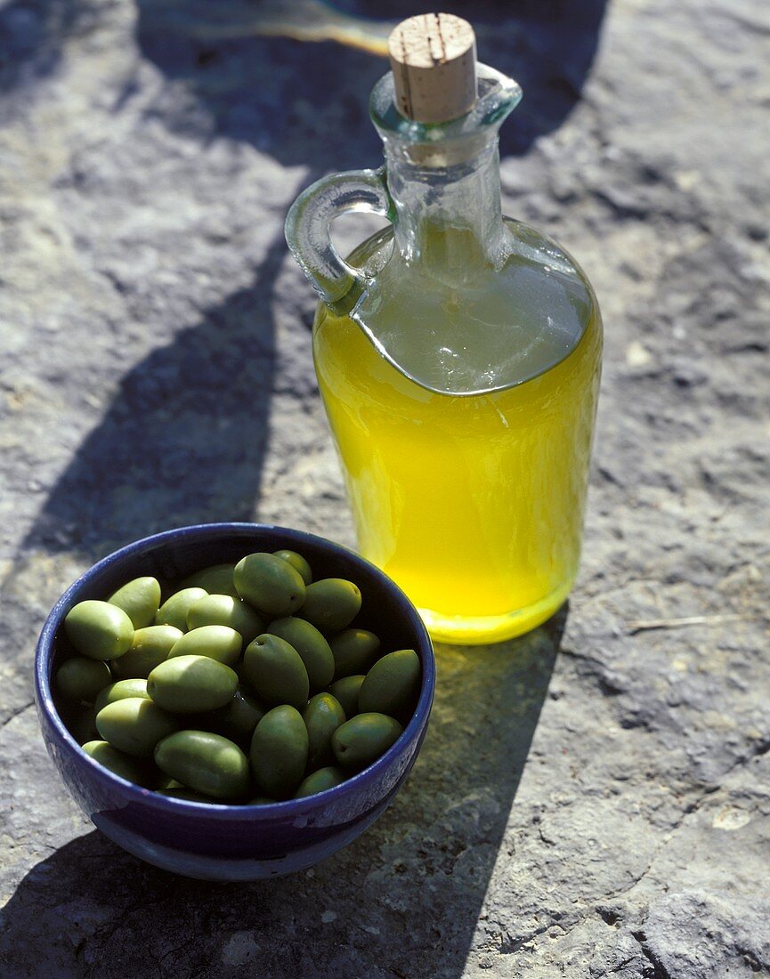 Olivenöl und eine Schale grüne Oliven auf Steinuntergrund