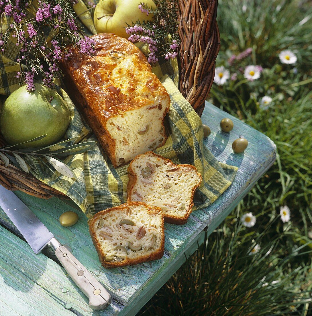 Oliven-Schinken-Kuchen, Äpfel und Kräuter im Korb auf Wiese