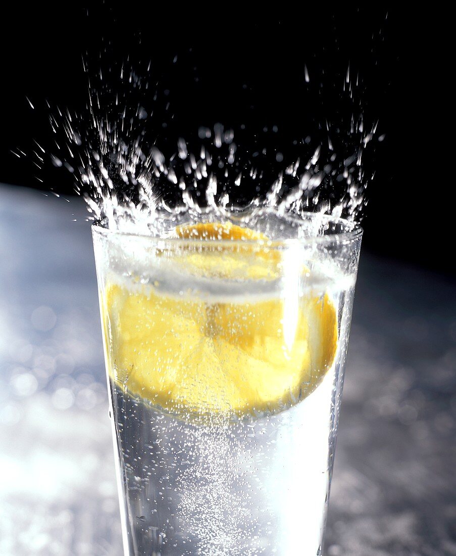 Zitronenscheibe fällt spritzend in Glas Mineralwasser