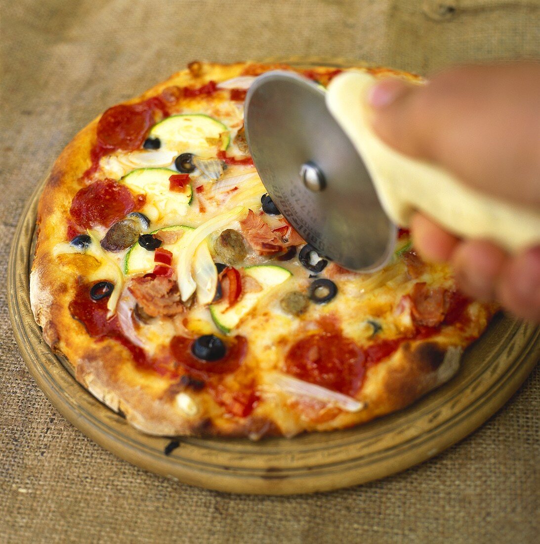 Gemüsepizza mit Schinken; Hand mit Pizzaschneider