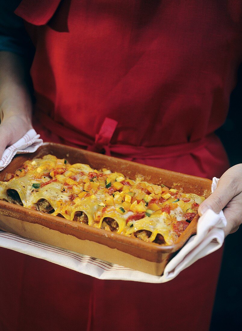 Woman in red apron holding baking dish of cannelloni