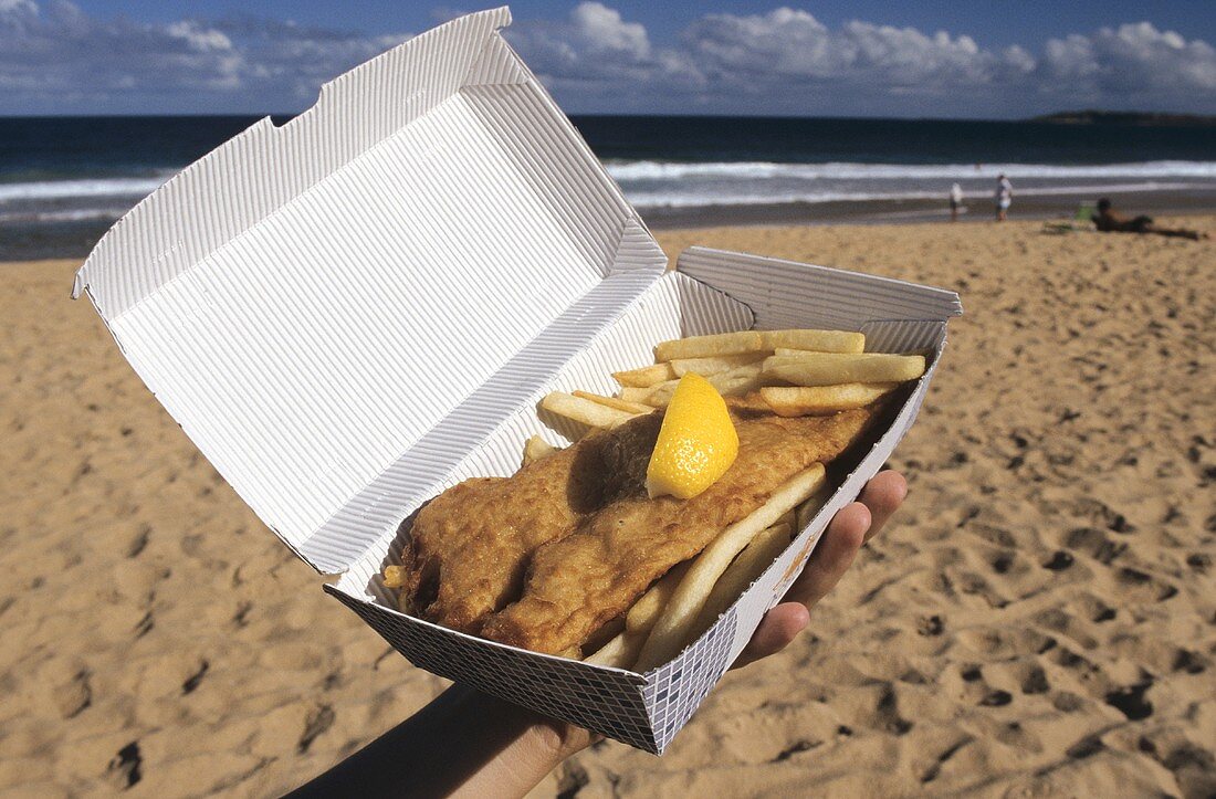 Hand hält Fish and Chips in einer Schachtel am Meer