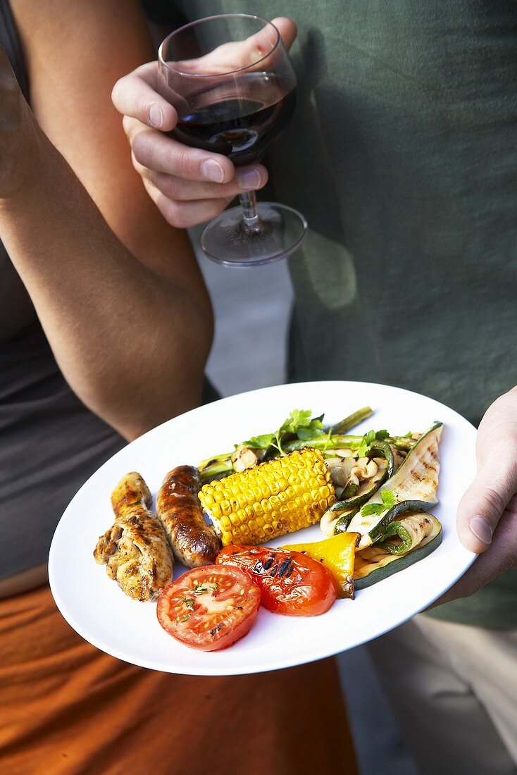 Hand holding plate of barbecued sausages, chicken & vegetables