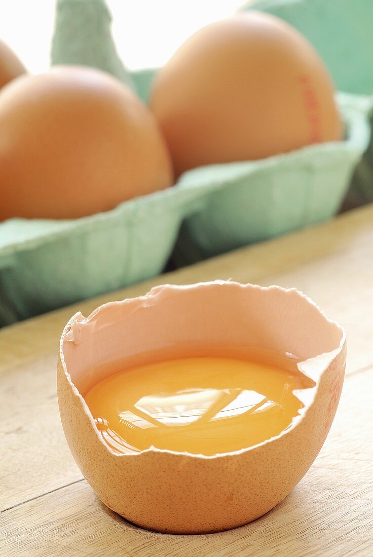 Aufgeschlagenes Ei, dahinter Eier im Eierkarton