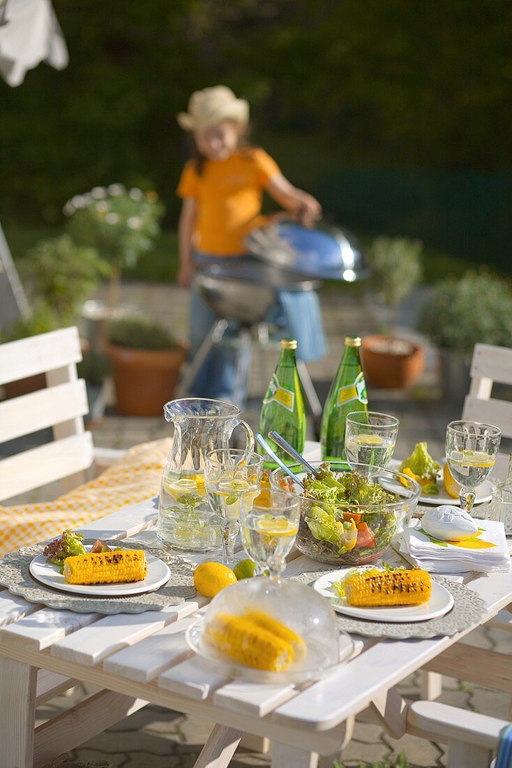 Gegrillte Maiskolben und Salat auf Tisch, Kind im Hintergrund