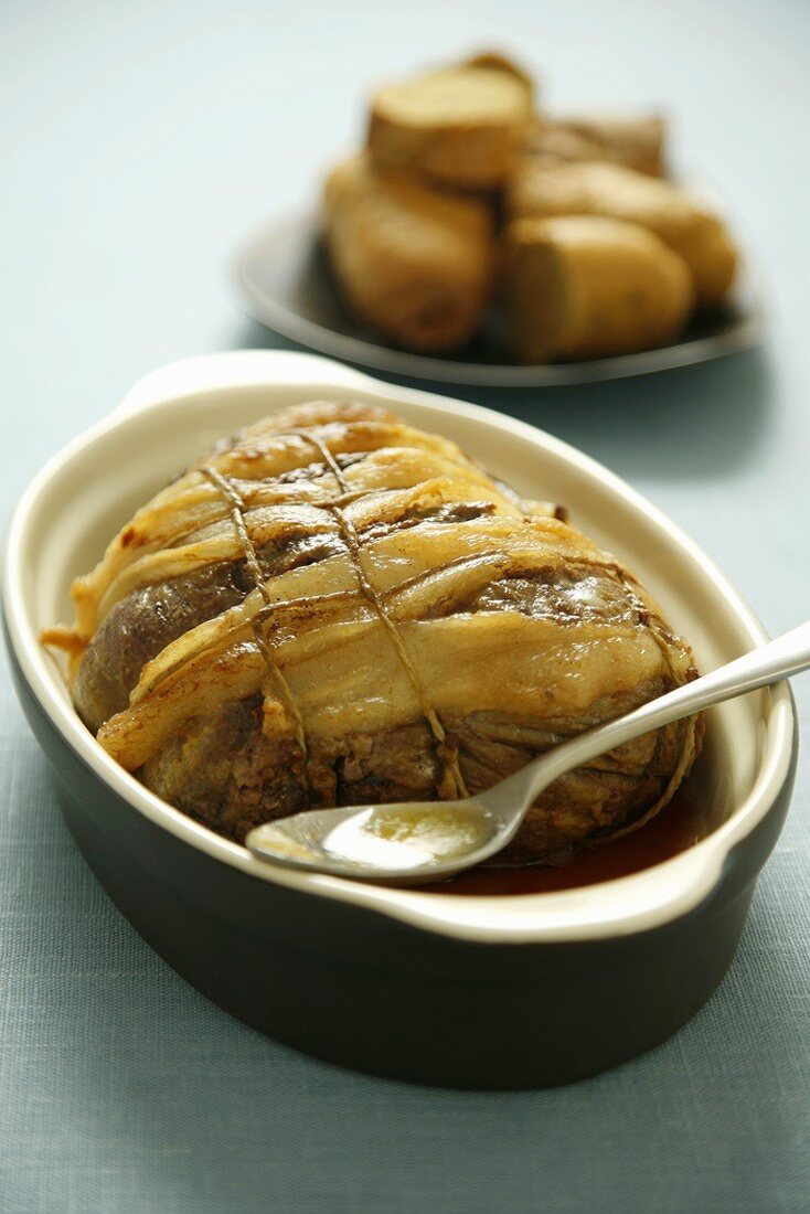 Roast beef with fat in roasting dish, potatoes in background