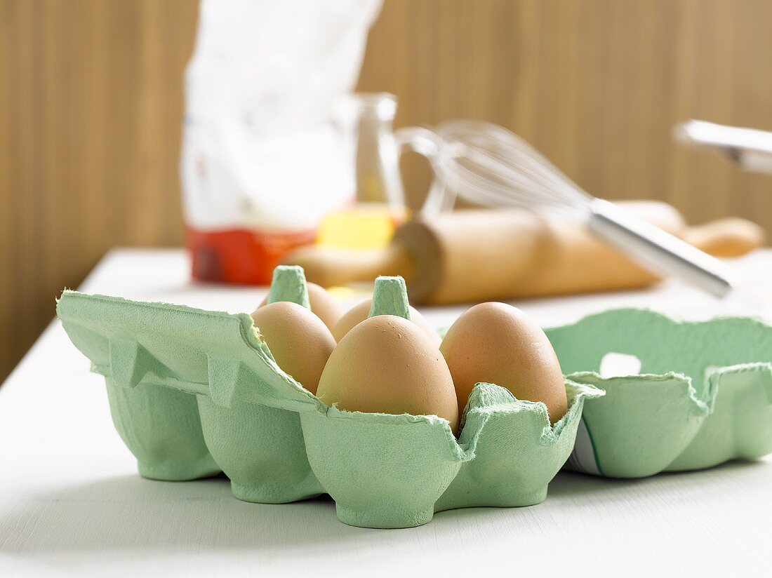 Eggs in egg box, rolling pin, whisk and flour behind