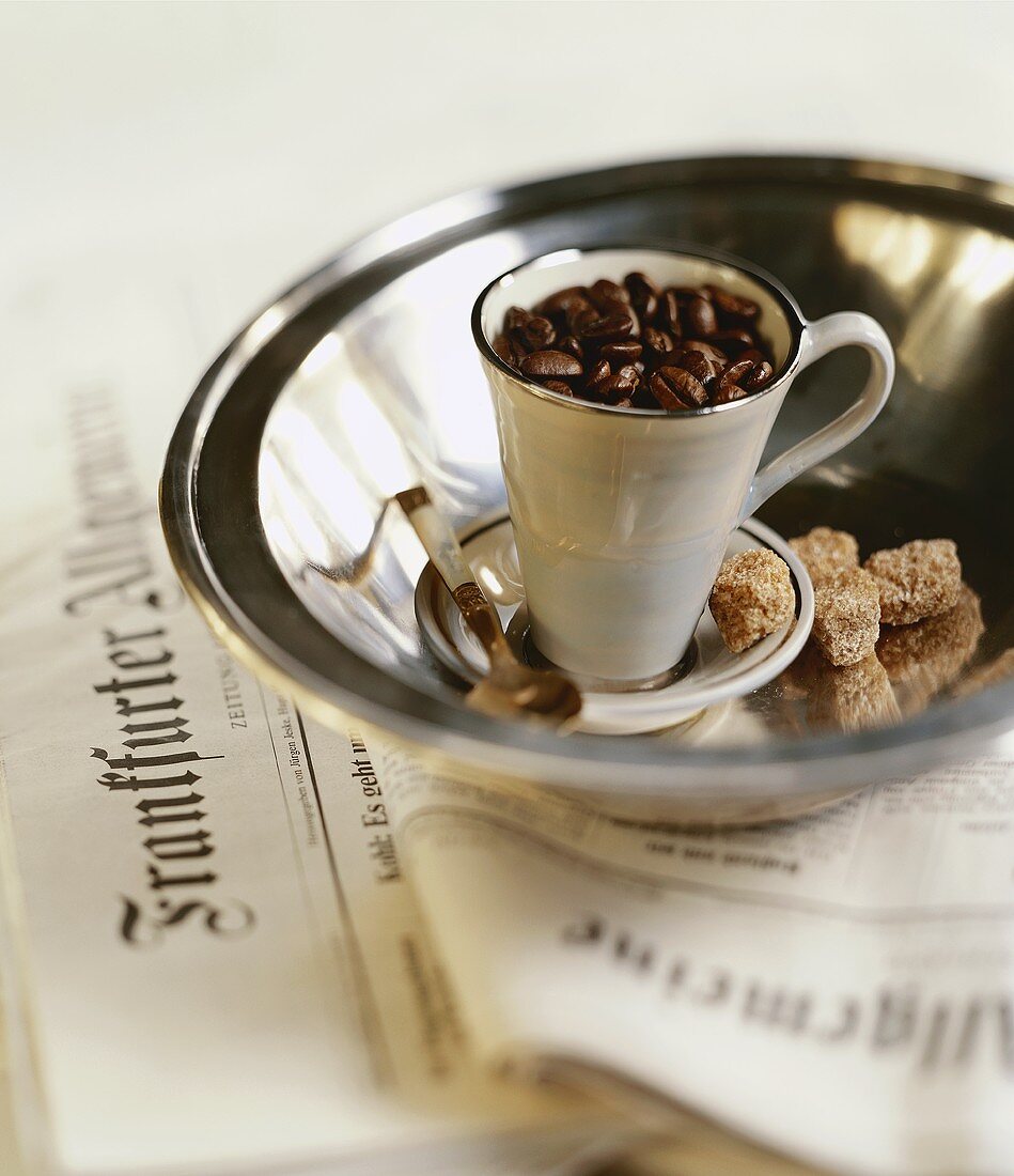 Coffee cup filled with coffee beans in silver bowl on newspaper