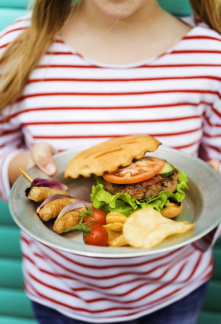 Woman holding plate of barbecued food: hamburger, sausages, crisps