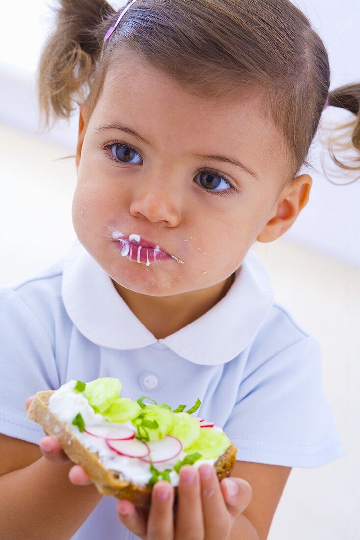 Little girl eating slice of bread with quark, cucumber and radishes