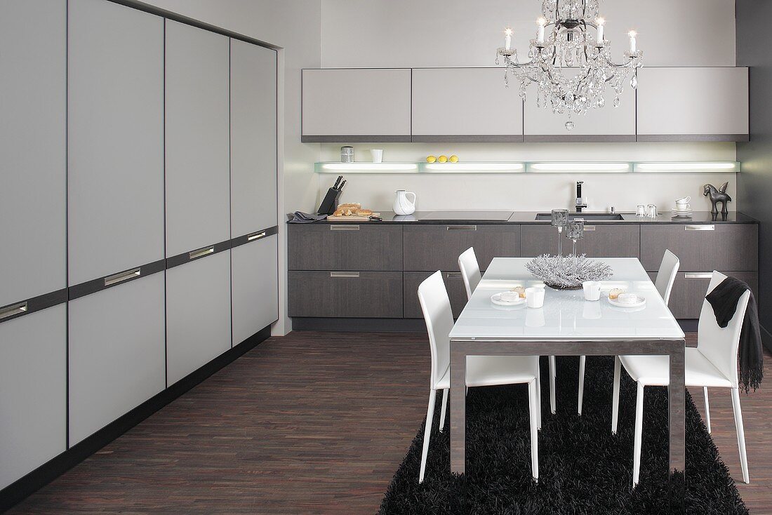 A stainless steel dining table with a white glass platter and white leather chairs under a chandelier in a modern kitchen with fitted cupboards