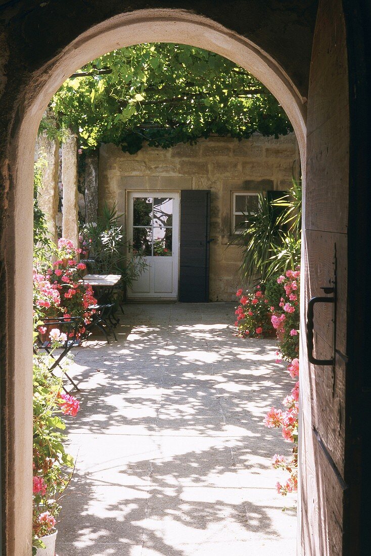 View through archway of sunny courtyard with shady, vine-covered pergola and abundant flowers