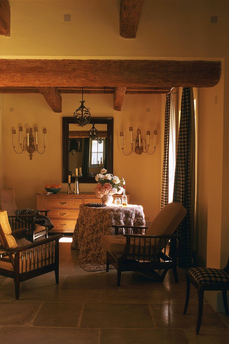 Schattiger Wohnraum mit Kommode, gerahmten Spiegel und Holzstühlen mit weichen Polstern; dazu ein runder Tisch mit bodenlanger Tischdecke und einem barocken Blumenstrauss