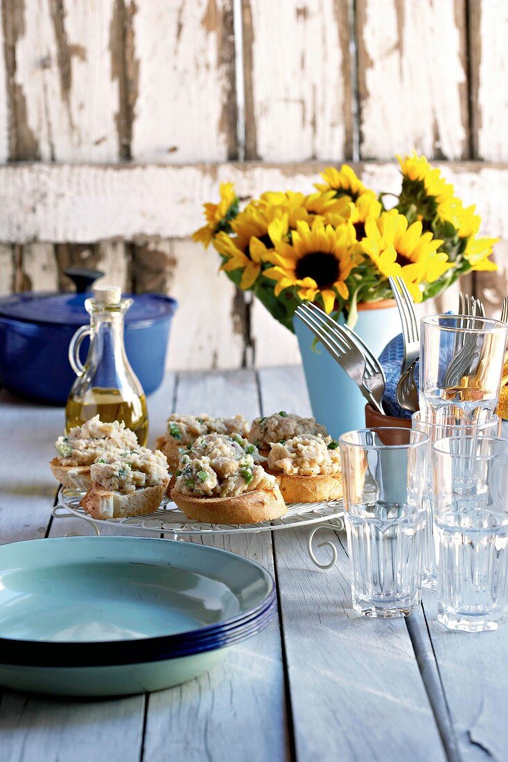 Crostini mit Bohnenpaste, Gläser, Sonnenblumenstrauss auf einem Tisch im Freien