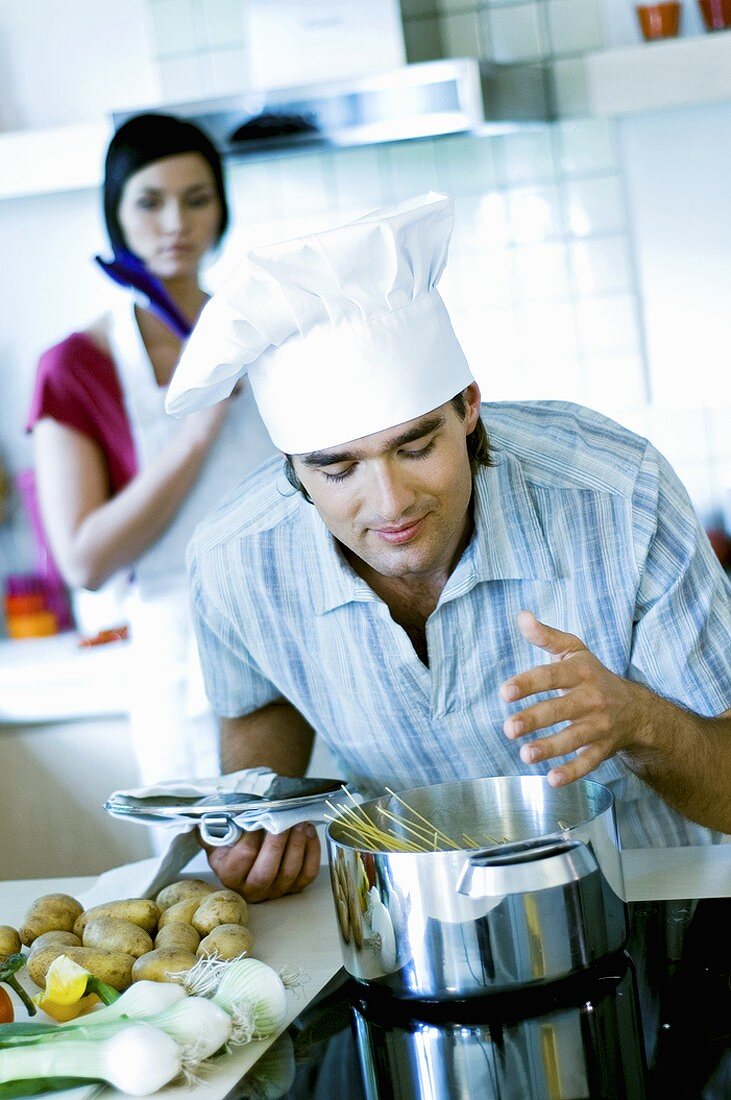 Man in chef's hat looking into pan, woman in background