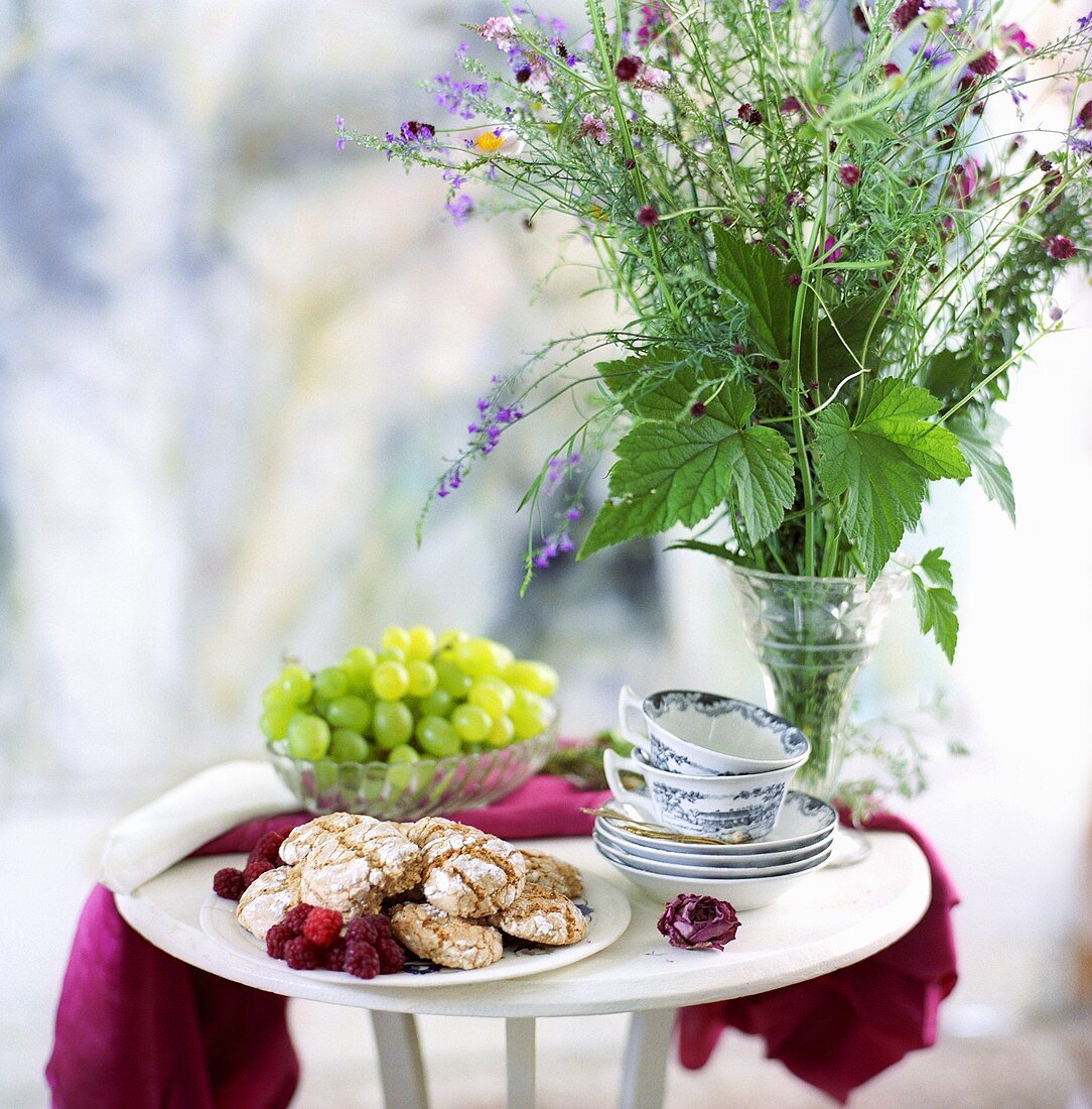 Almond biscuits, raspberries, grapes, cups & saucers on a table