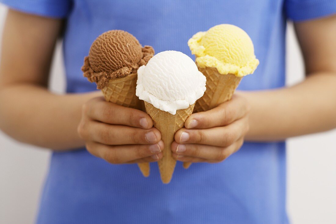 Hands holding three ice cream cones: chocolate, coconut, vanilla