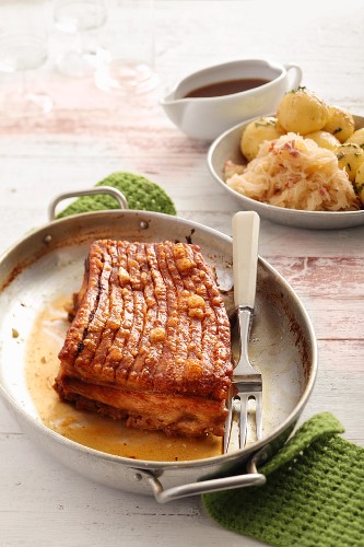 Knusprig gebratener Schweinebauch am Stück, im Hintergrund Sauerkraut und Kartoffelknödel