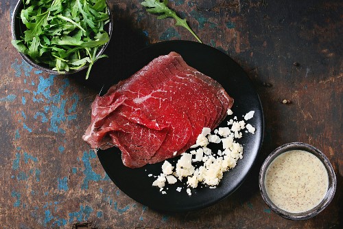 Ingredients for carpaccio: Raw sliced beef, mustard and parmesan sauce, cheese, bowl of arugula