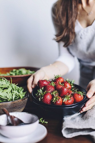 A woman is placing a bowl of strawberries on a table, getting ready to put together a strawberry and spinach salad
