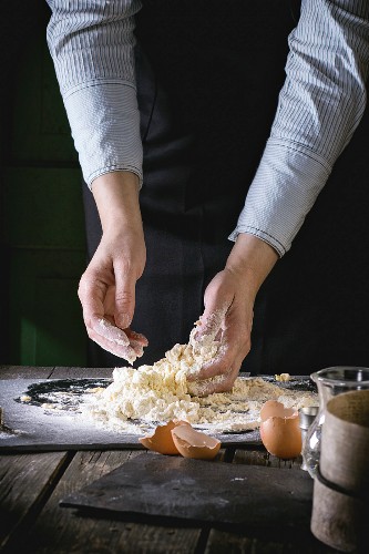 Female hands knead the dough for pasta on old wooden kitchen table, broken eggs at foreground