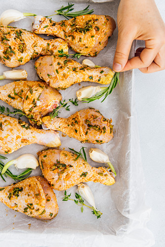 Woman placing rosemary sprigs among chicken drumsticks in a baking sheet