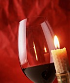 A red wine glass next to a burning candle in front of a red background