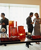 Decorative red glass vases and gerberas on a tray arranged on a table with party guests in the background