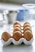 Frische Eier im Eierkarton, im Hintergrund Backutensilien