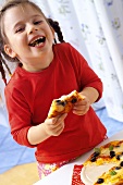Kleines Mädchen isst lachend ein Stück Gemüsepizza