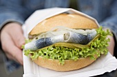 Hands holding herring in bread roll on paper plate