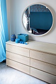 Light wood chest of drawers and turquoise accessories below round mirror on wall
