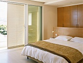 Doppelbett mit Raumteiler-Kopfteil in modernem Schlafraum mit Fensterläden vor breiten Schiebefenstern